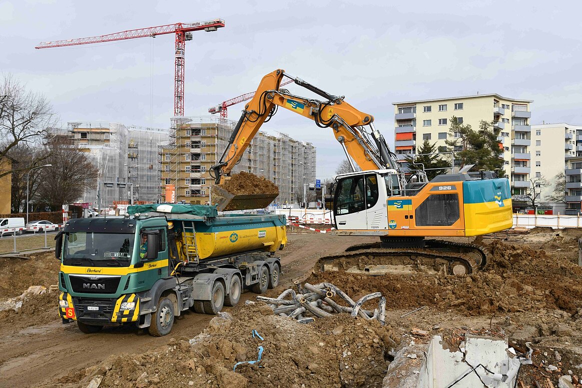 Ein Bagger belädt einen Lastwagen mit Aushub einer Baustelle.